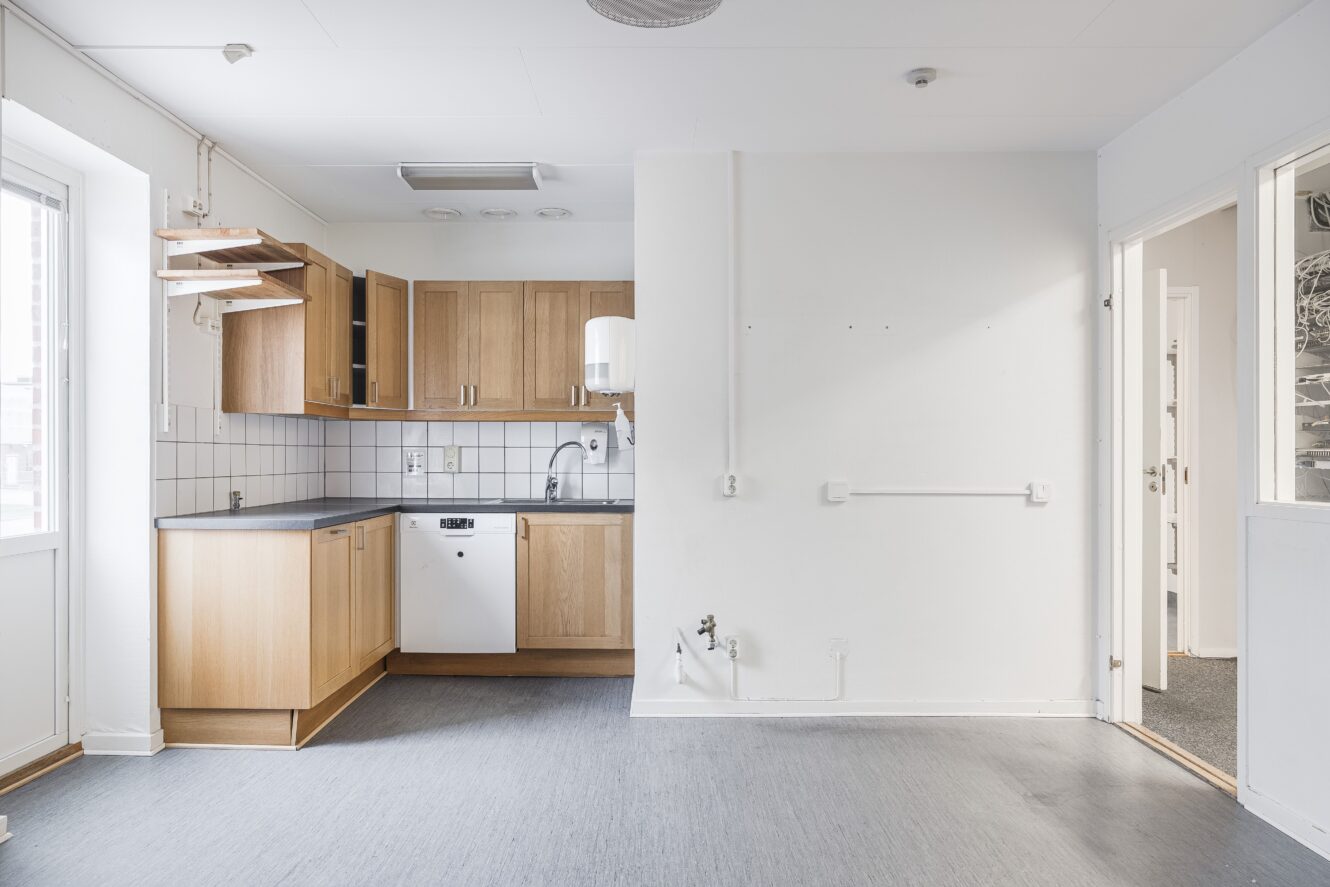 Ett litet, omöblerat kök med skåp i trä, vita kakelplattor, diskmaskin och intilliggande tomt utrymme i ett ljust, minimalistiskt rum.