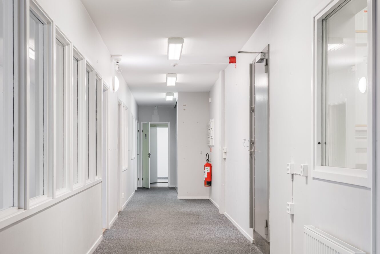 En ren, väl upplyst korridor med vita väggar, flera fönster till vänster, en brandsläckare på den högra väggen och stängda dörrar i slutet.