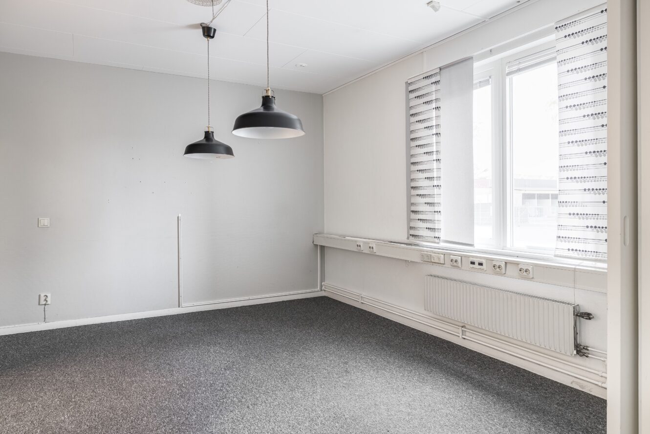 Tomt kontorsrum med grå heltäckningsmatta, vita väggar, två svarta pendellampor, ett stort fönster med mönstrade persienner och flera eluttag längs väggen.