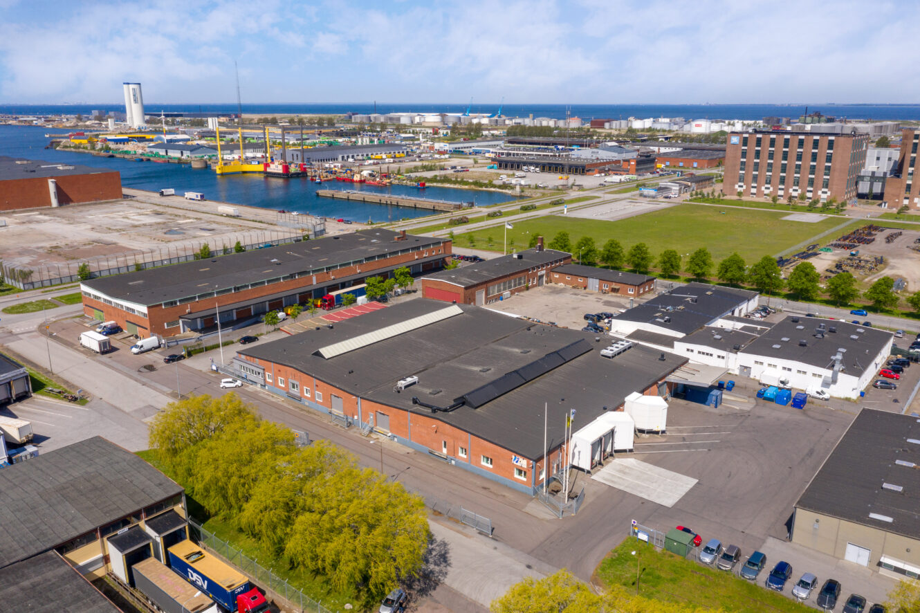 Flygfoto över ett industriområde med lagerlokaler, kontorsbyggnader, parkerade fordon, grönområden och en hamn med fartyg i bakgrunden.