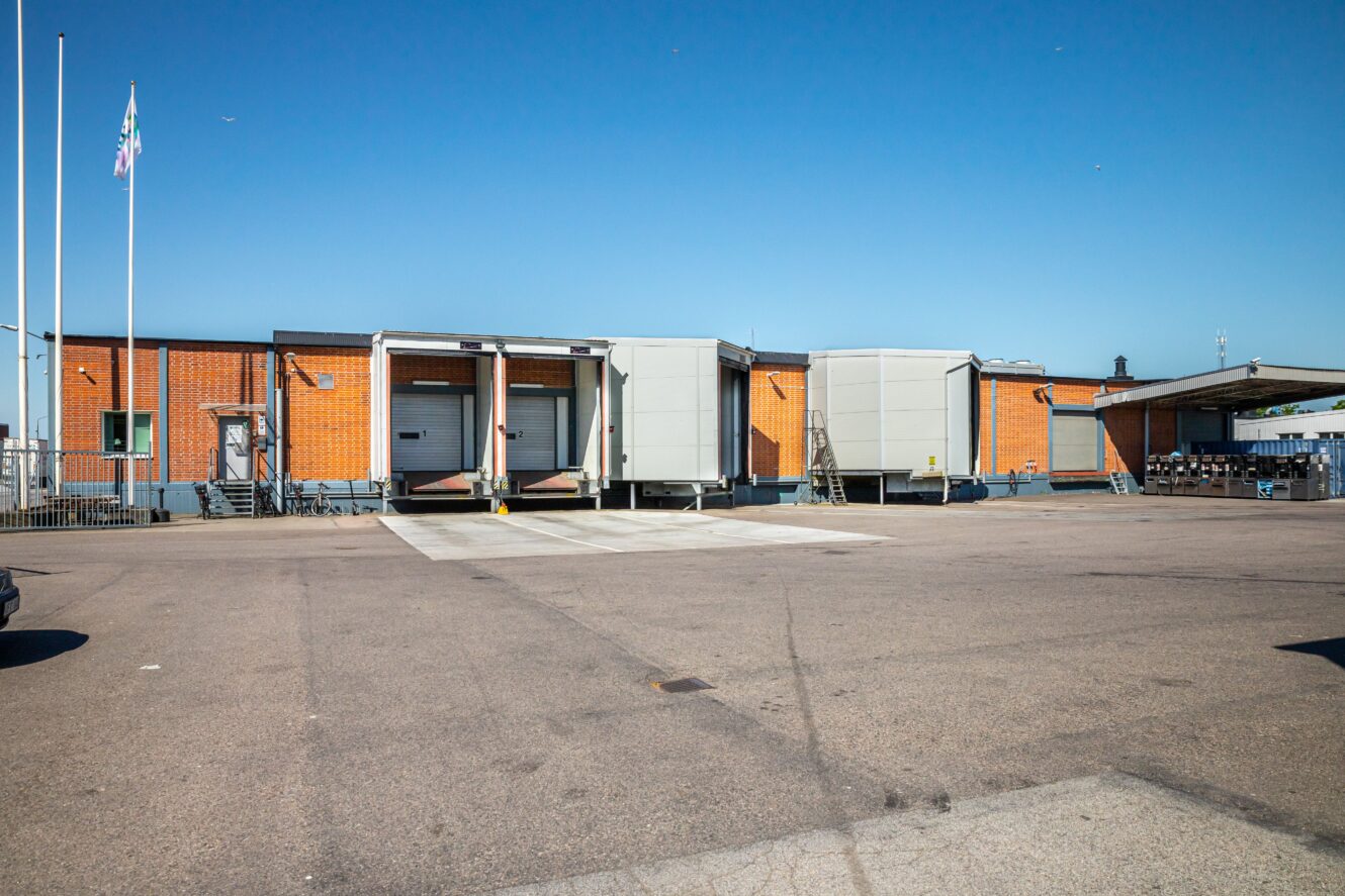 Ett distributionslager med tre lastbryggor, tegelväggar och en klarblå himmel, sett från en tom asfalterad tomt.