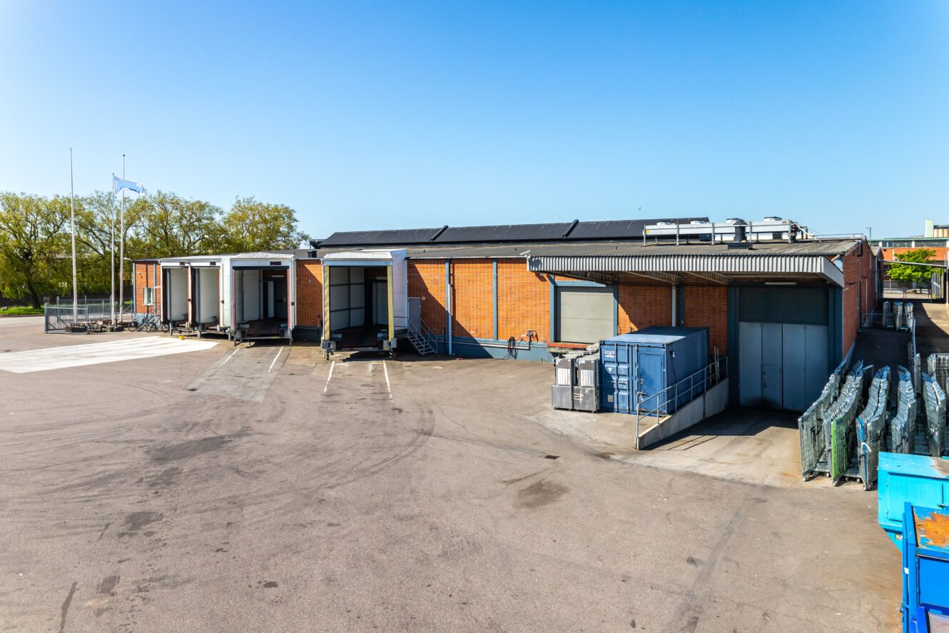 Ett industriellt tegelmagasin med flera lastkajer, metallcontainrar och en öppen asfalterad tomt under en klarblå himmel.