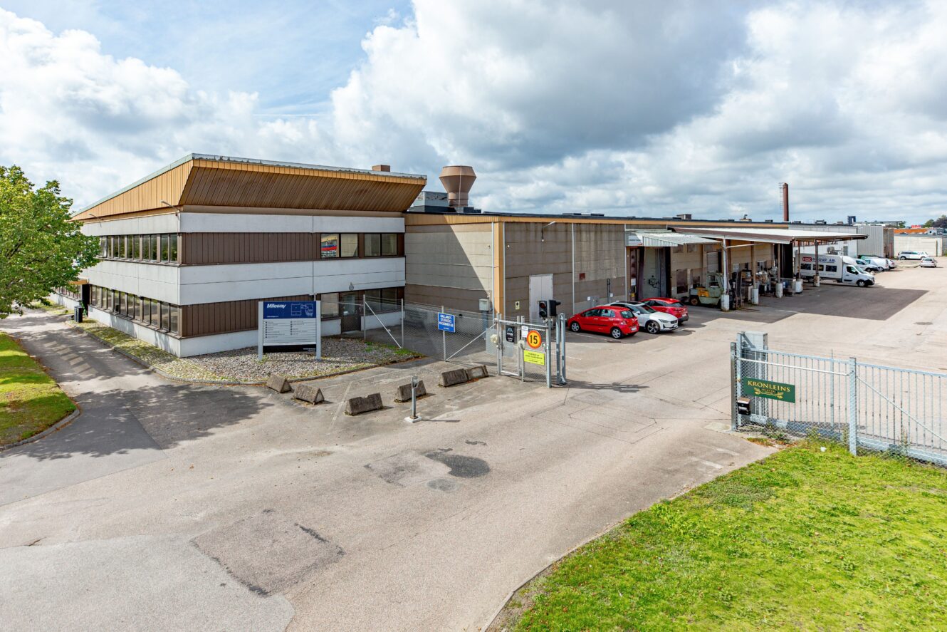 En stor kommersiell eller industriell byggnad med flera garageplatser, parkerade fordon och en inhägnad entré, under en delvis molnig himmel.
