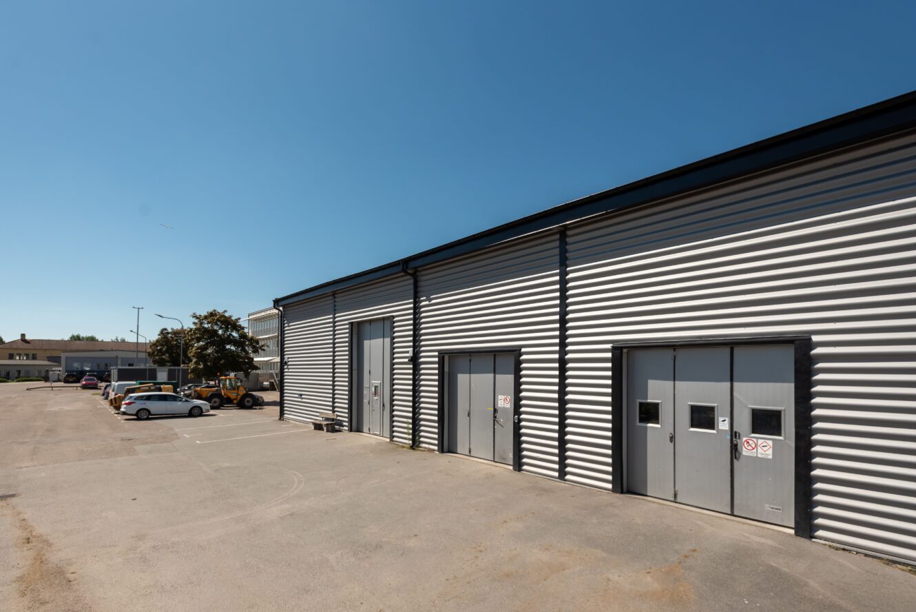 Ett modernt industriellt lager med korrugerad metallfasad, tre stora dörrar och en asfalterad parkeringsplats med flera parkerade bilar under en klarblå himmel.