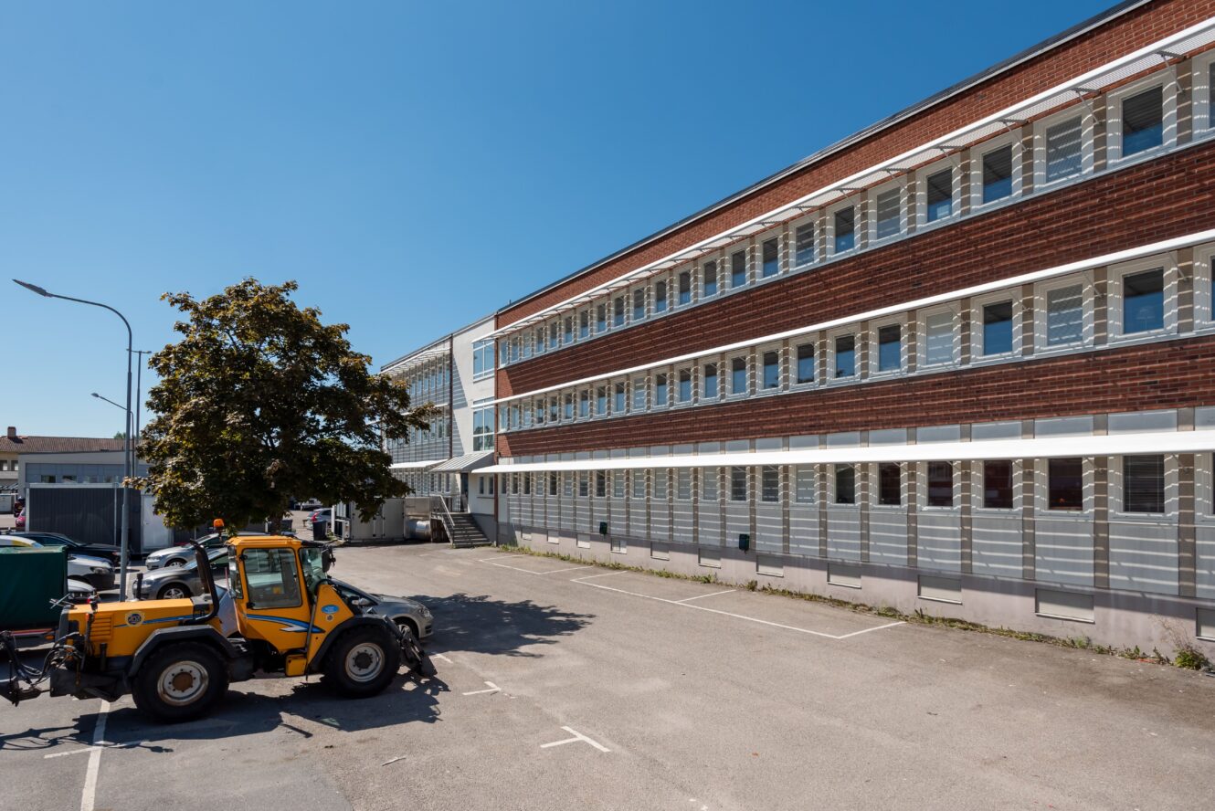 En gul byggbil står parkerad på en tom parkeringsplats bredvid en stor trevåningsbyggnad i tegel och vitt under en klarblå himmel.