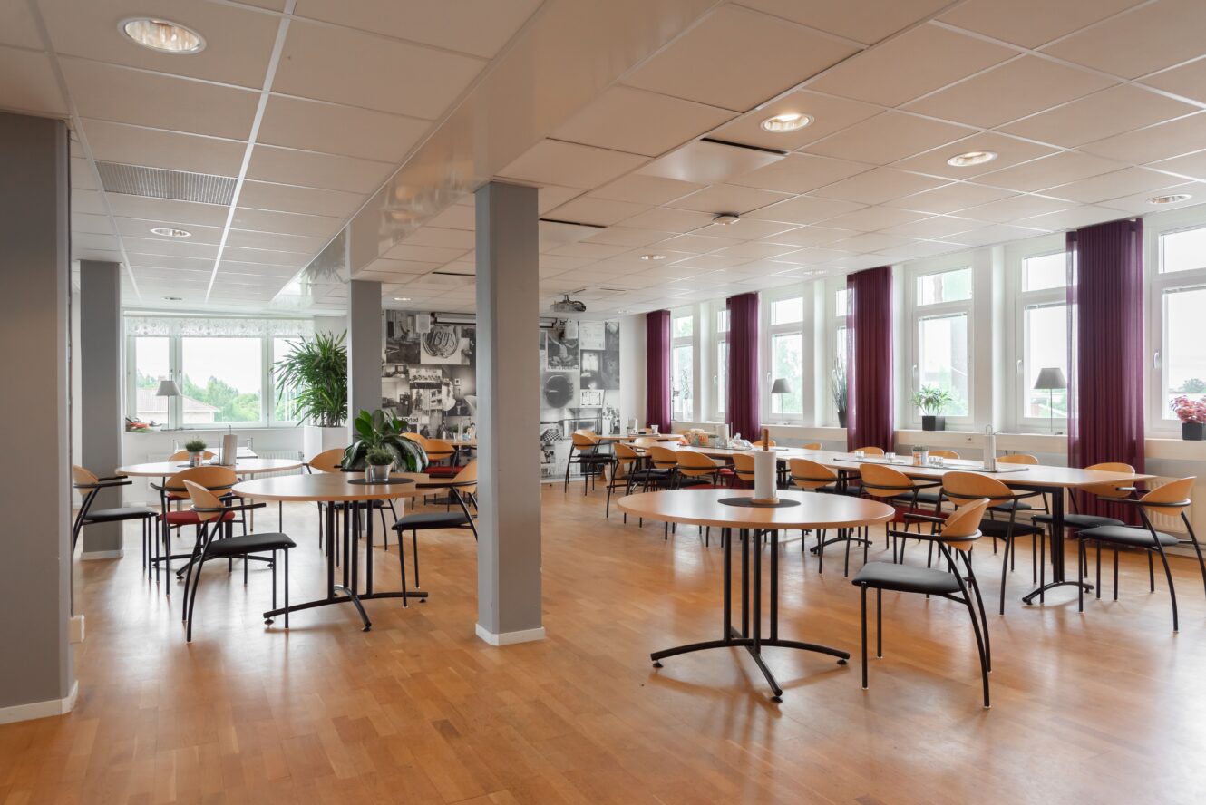 En rymlig och modern cafeteria med trägolv, runda bord och prydligt placerade stolar. Stora fönster släpper in naturligt ljus; krukväxter och väggkonst syns.