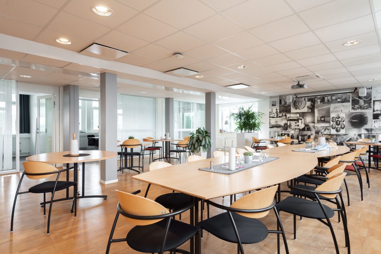 Modernt fikarum på kontor med bord i ljust trä, svarta stolar, krukväxter och stora fönster. En vägg pryds av en svartvit fototapet. Rummet är snyggt och väl upplyst.