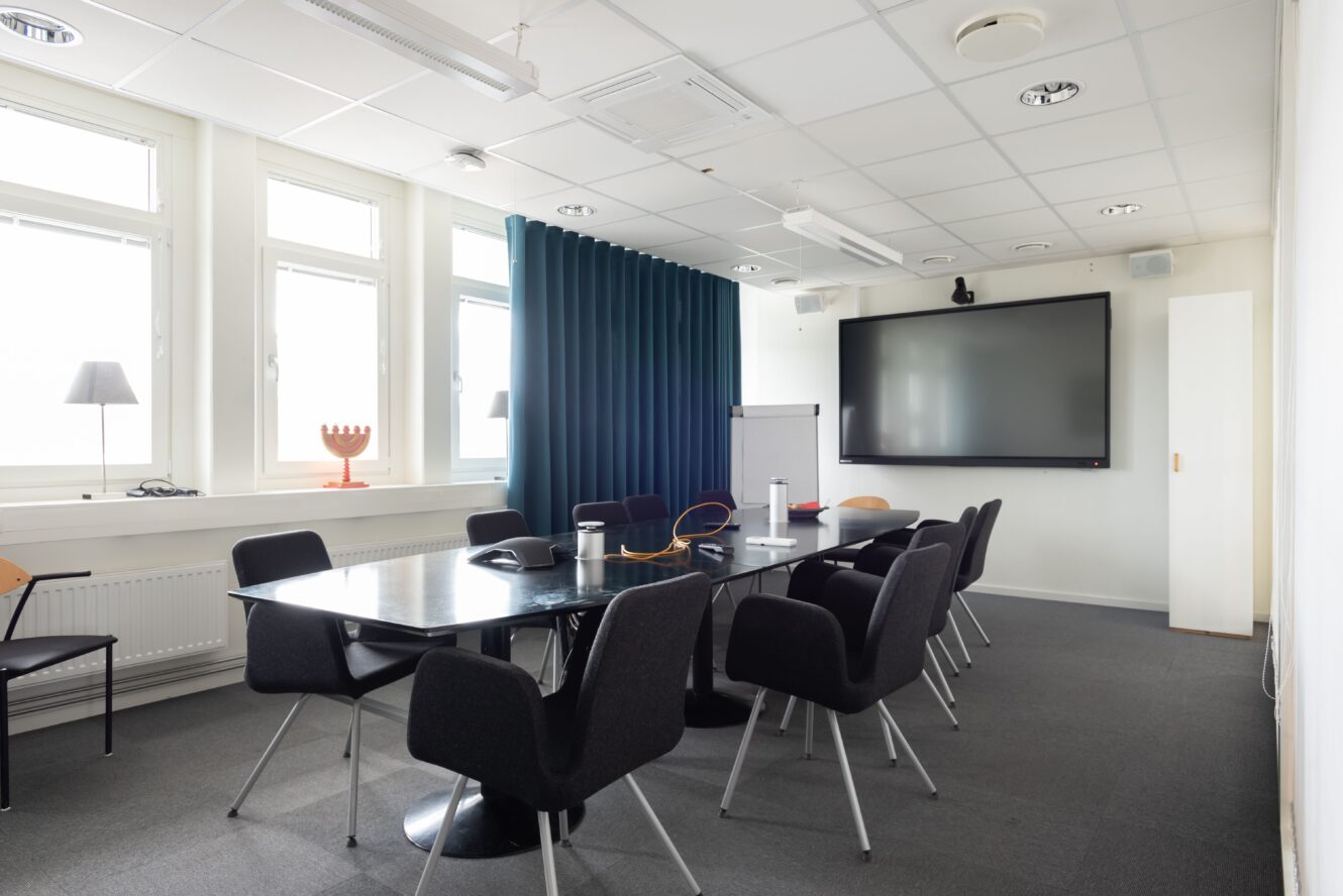 Ett modernt konferensrum med ett stort svart bord, åtta svarta stolar, en väggmonterad skärm, ett blädderblock och fönster som släpper in naturligt ljus.