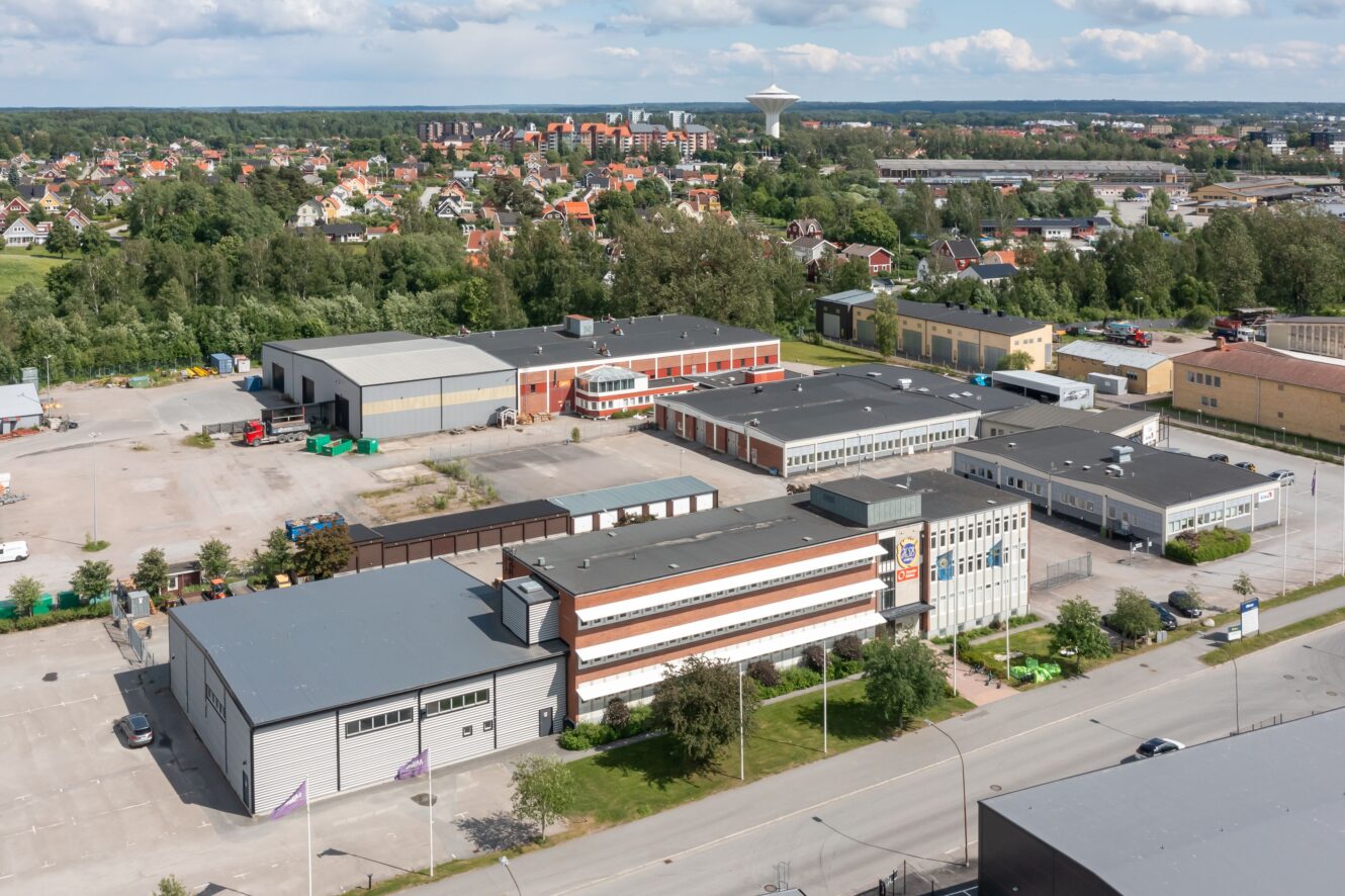 Flygfoto över ett industrikomplex med flera lager- och kontorsbyggnader, beläget nära en huvudväg, med ett bostadsområde och grönska i bakgrunden.