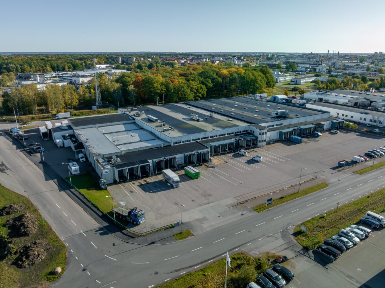 Flygbild över ett stort industrilager med lastkajer, lastbilar och en parkeringsplats, omgivet av vägar och träd, under en klar himmel.