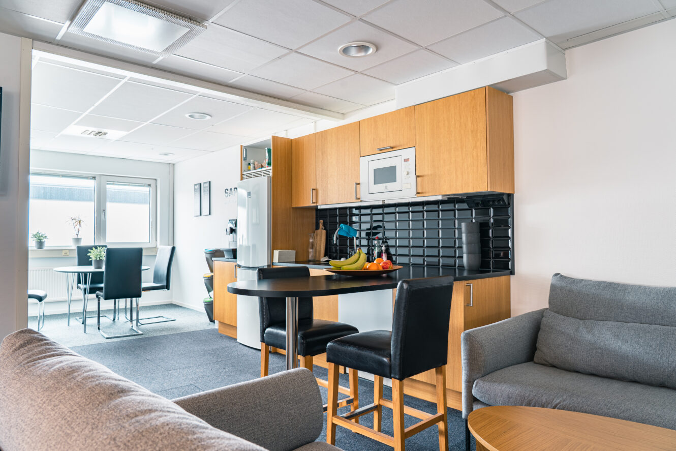 Modernt kök och vardagsrum med träskåp, svart stänkskydd, litet matbord, grå soffor och naturligt ljusinsläpp från stora fönster. Fruktskål och vitvaror synliga på bänkskivan.