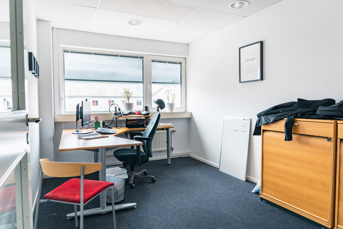 Ett litet kontor med ett skrivbord, dator, stol, två fönster, ett diplom på väggen, ett skåp, en whiteboardtavla och en jacka ovanpå skåpet.