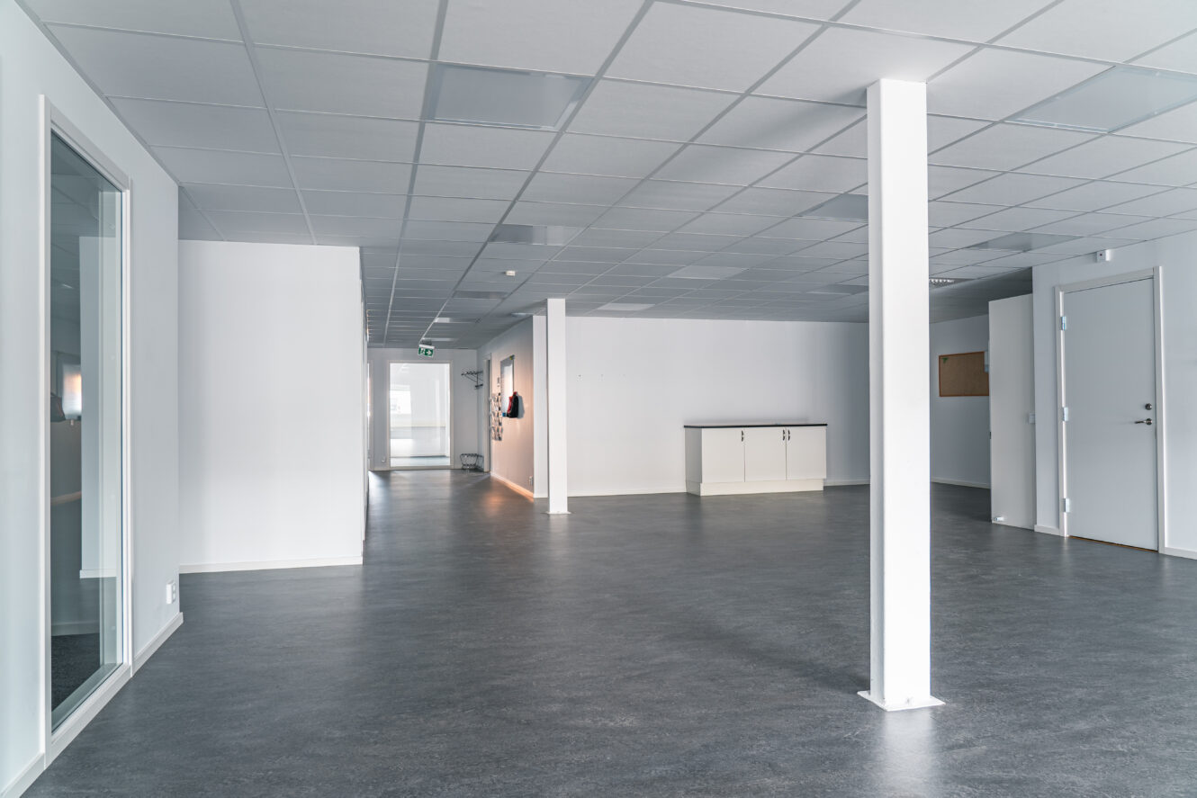 Tomt kontorsutrymme med vita väggar, glasparti till vänster, grått golv, takplattor, stödpelare och ett litet skåp mot bortre väggen.