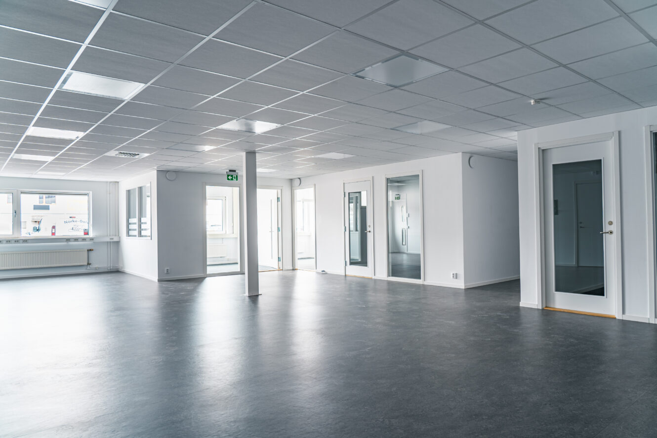 Tomt modernt kontorsutrymme med vita väggar, stora fönster, glasdörrar och ett grått golv, upplyst av taklampor och naturligt ljus.