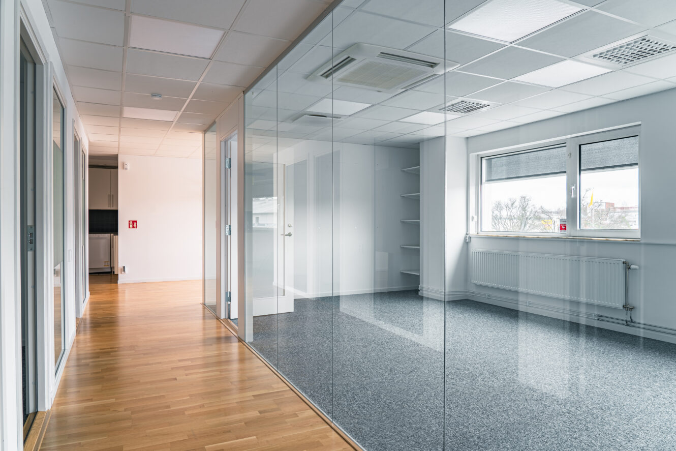 Moderna tomma kontorslokaler med glasväggar, trä- och heltäckningsmattor, stora fönster, vita väggar och inbyggda hyllor.