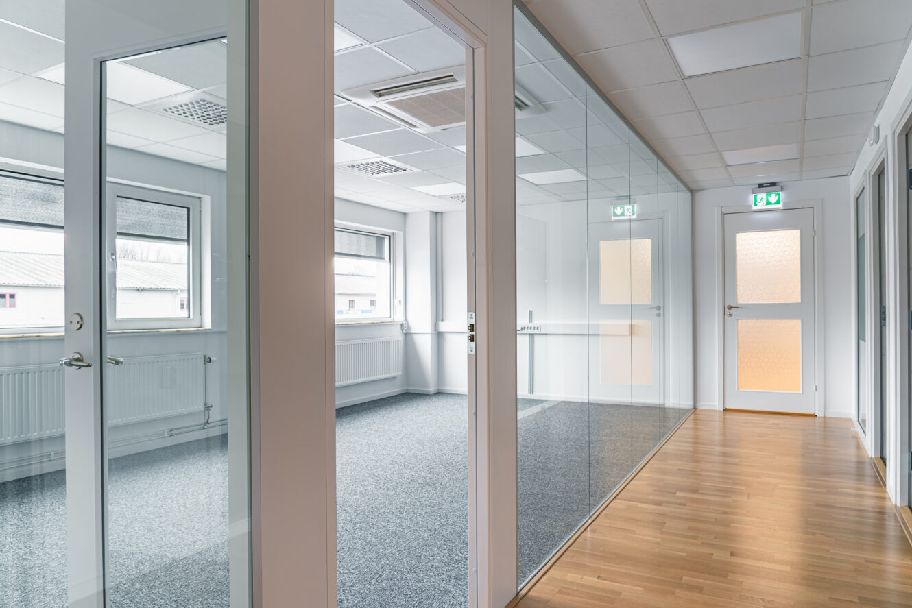 Modern kontorskorridor med glasväggar, stängda dörrar, heltäckningsmatta och trägolv samt stora fönster som släpper in naturligt ljus.
