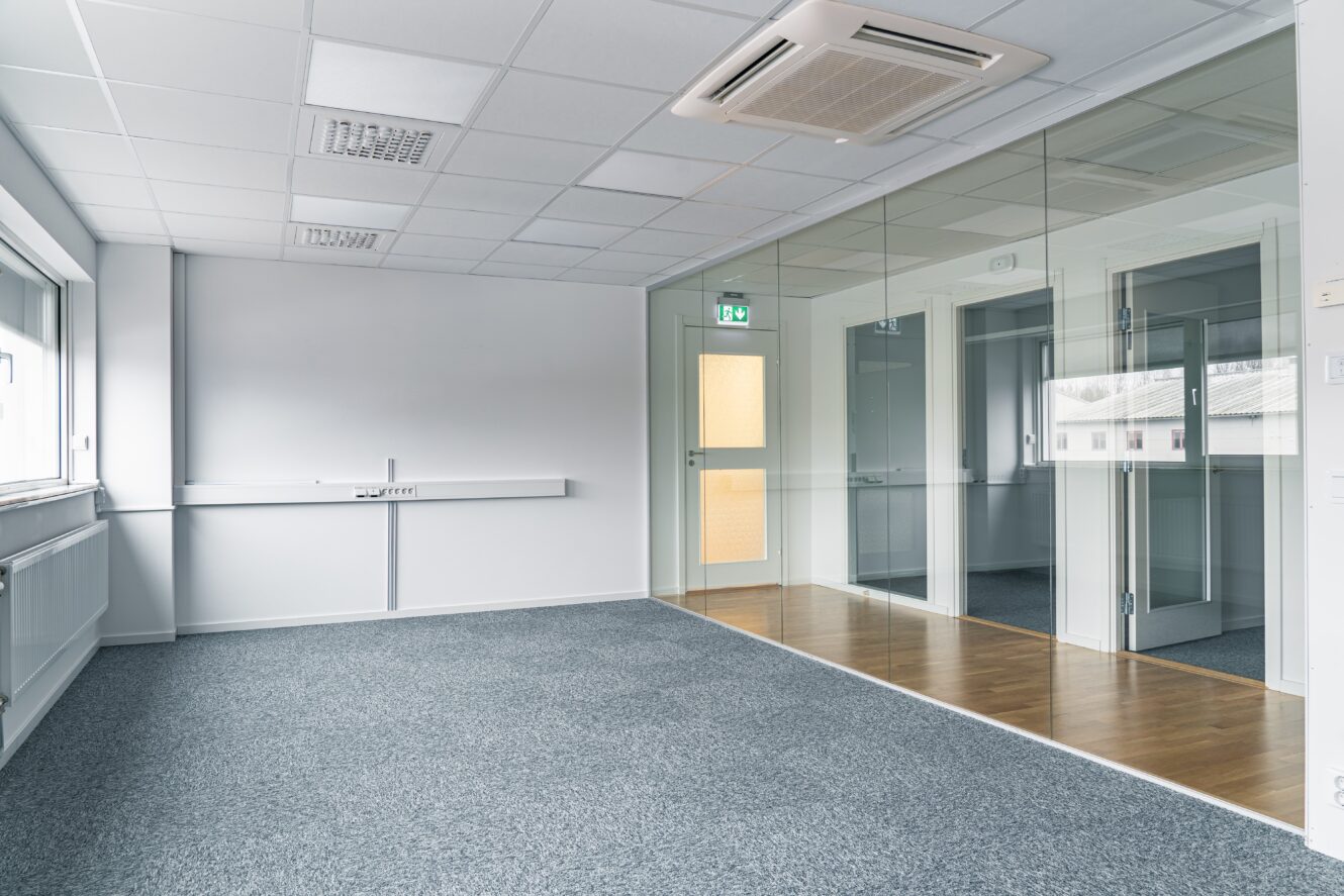 Tomt modernt kontorsutrymme med grå matta, vita väggar, glasvägg, taklampor och stora fönster som släpper in naturligt ljus.