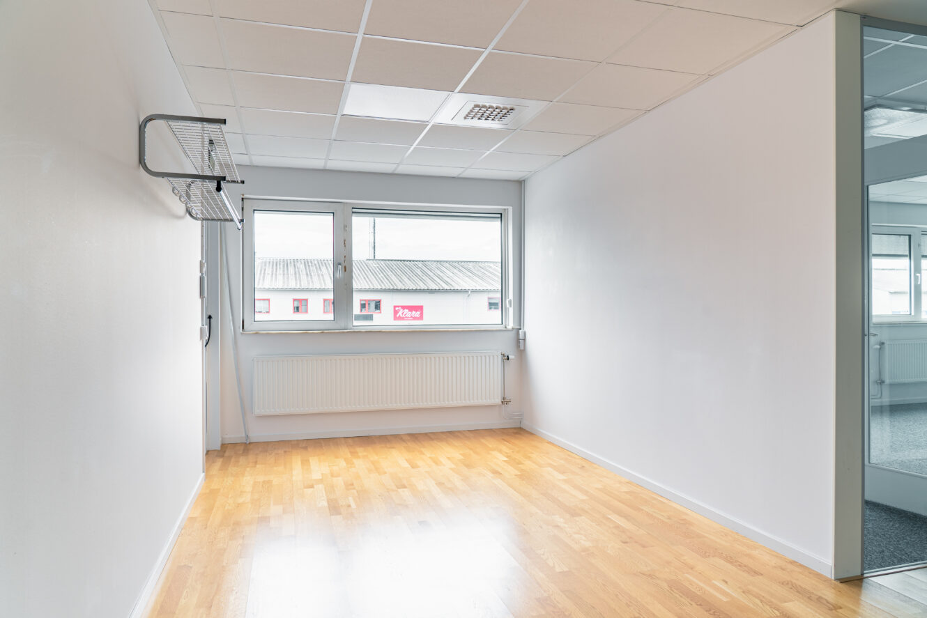 Tomt kontorsrum med vita väggar, takplattor, trägolv, fönster med radiator under, väggmonterad hylla och en glasdörr som leder till ett annat rum.