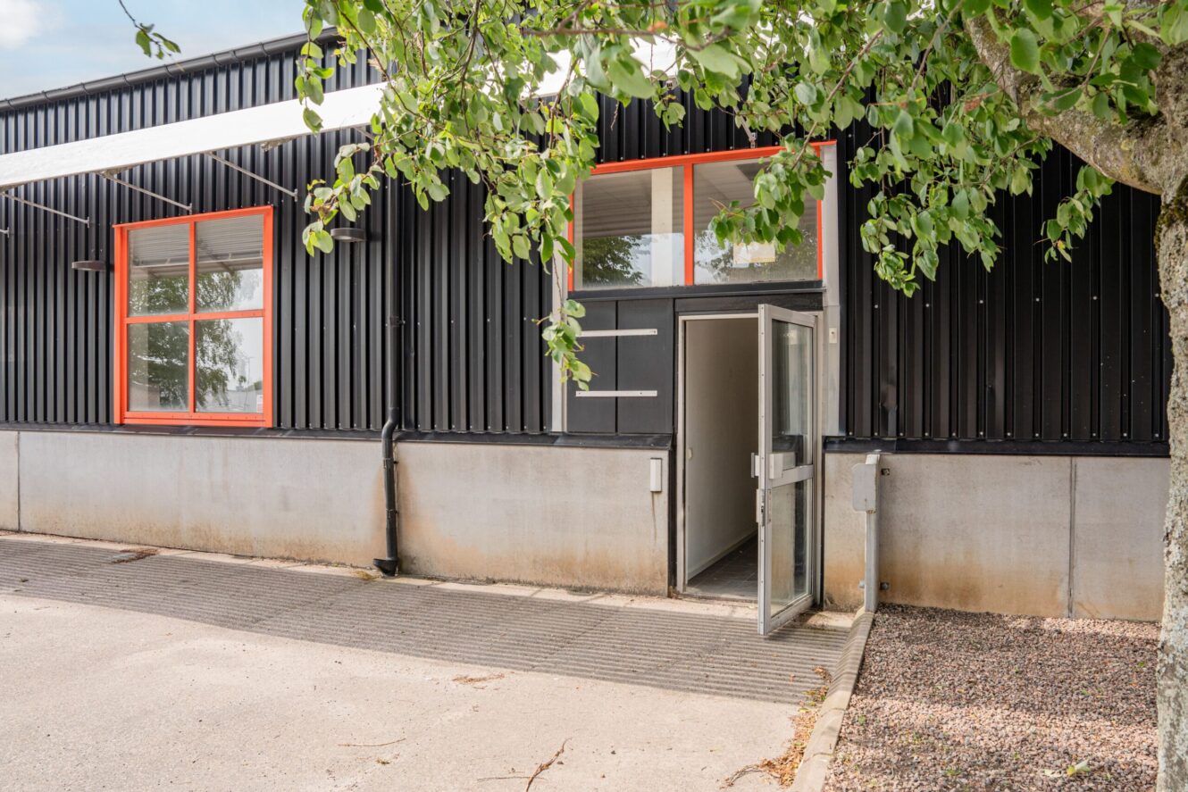 Envånings industribyggnad med svart metallbeklädnad, stora fönster med orange dekor och en öppen glasdörr som leder in. Träd och grus i förgrunden.
