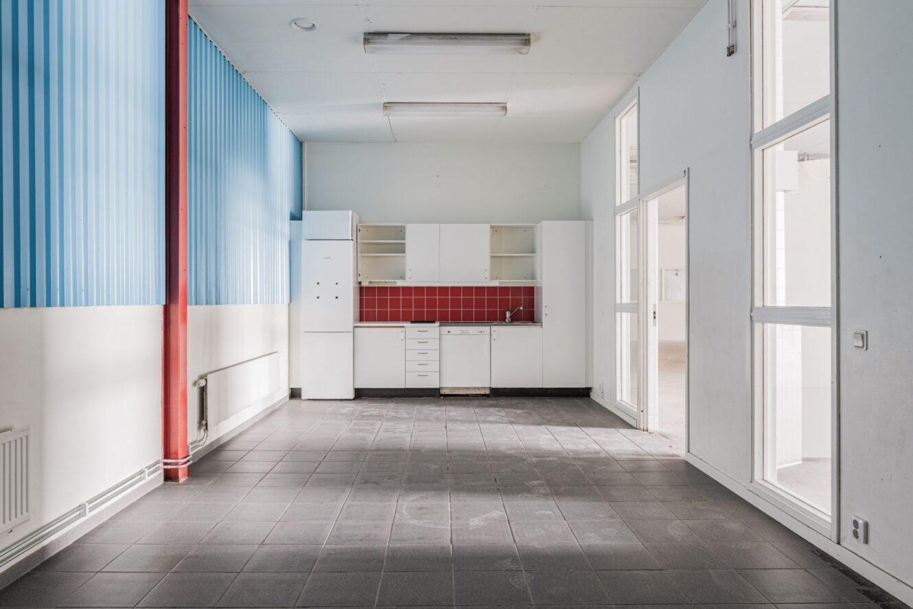 Ett litet, tomt kök med vita skåp, rött stänkskydd och grått klinkergolv, beläget i ett rum med högt i tak och blåvita väggar.