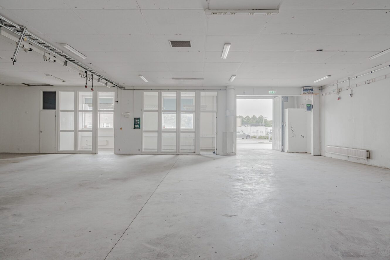 Tomt industrirum med vita väggar, stora fönster, lysrör i taket, synliga armaturer och öppna dubbeldörrar som leder ut.