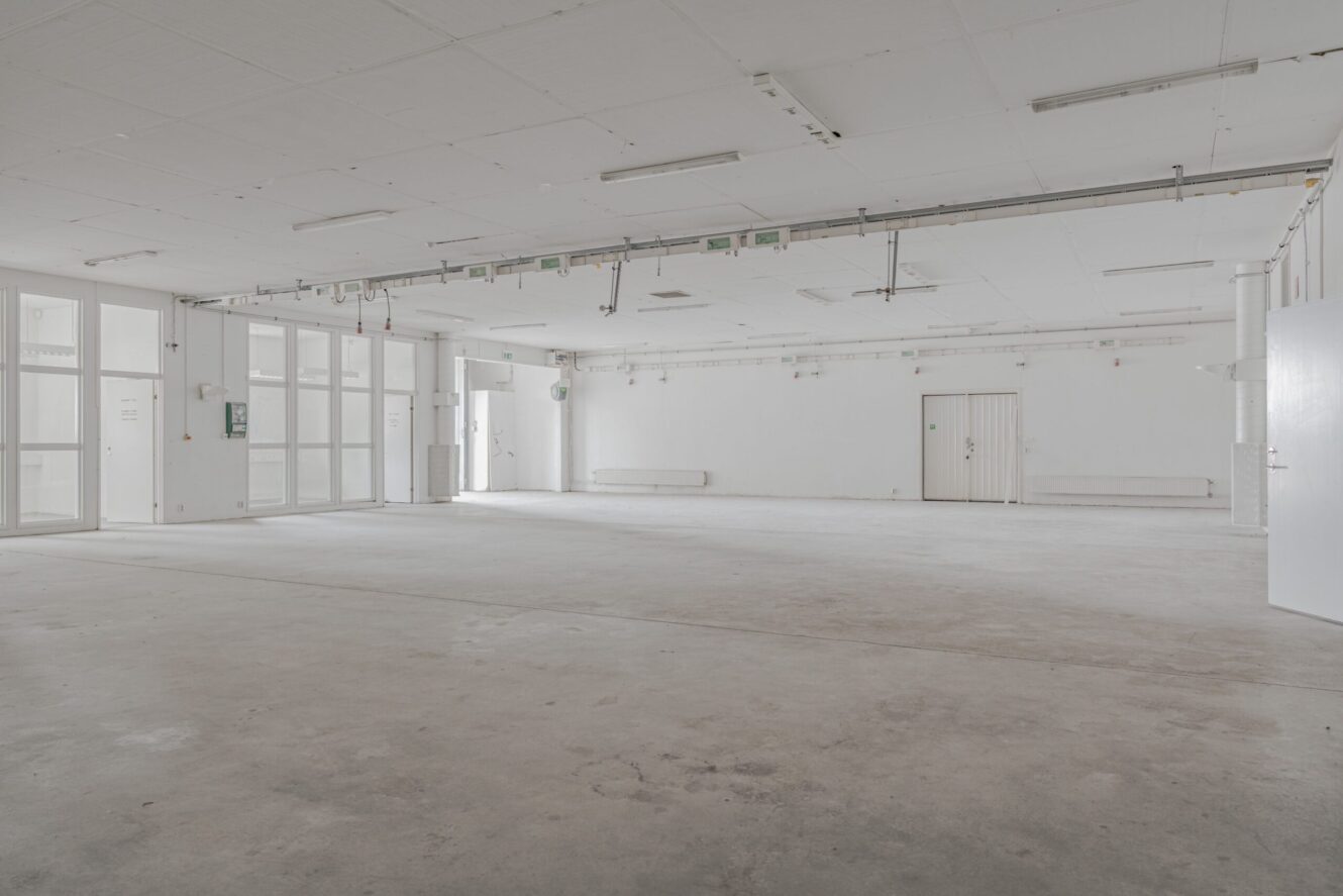 Stort tomt industrirum med vita väggar, betonggolv, lysrör och flera dörrar och fönster längs väggarna.