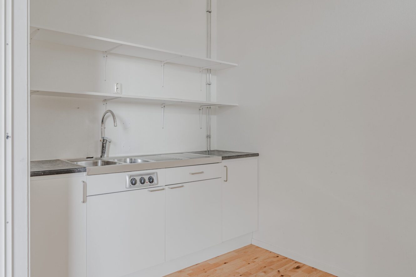 Ett litet, minimalistiskt kök med vita skåp, en diskho i rostfritt stål, öppna hyllor och trägolv mot en vit vägg.