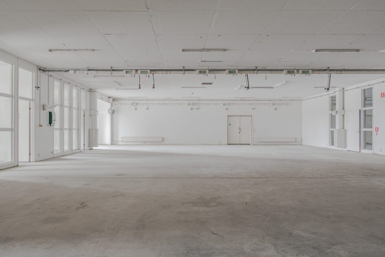 Stort, tomt industrirum med vita väggar, betonggolv, taklampor och stora fönster på vänster sida.