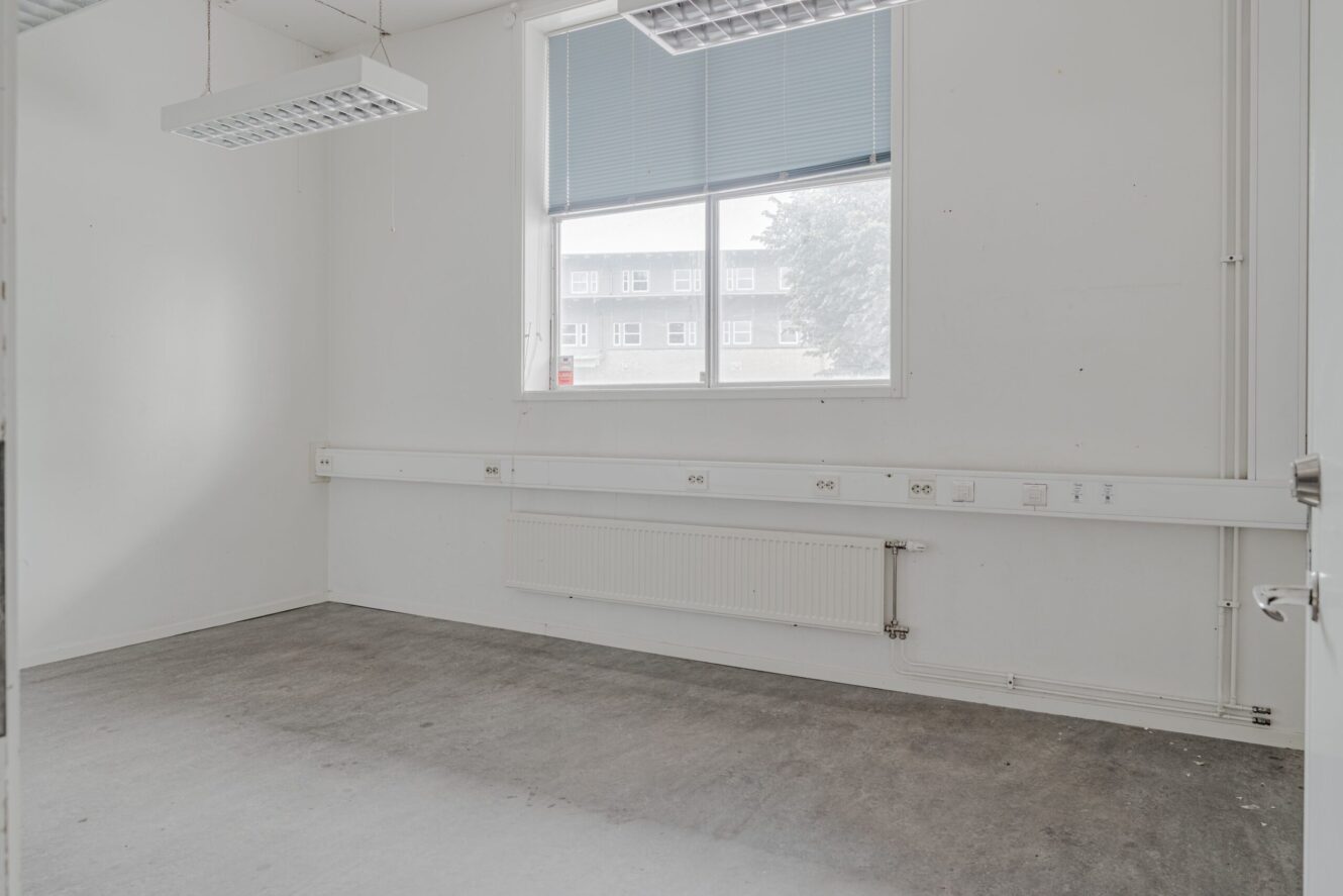 Tomt kontorsrum med vita väggar, ett stort fönster, grått golv, lysrör i taket och eluttag längs väggen.
