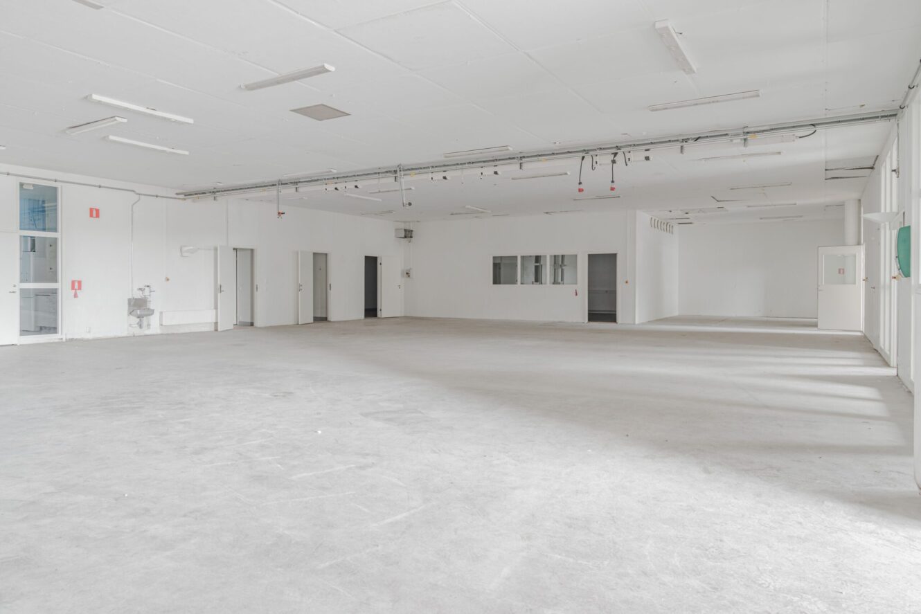 Stort, tomt industriellt eller kommersiellt utrymme med vita väggar, flera dörrar, taklampor och några väggmonterade armaturer synliga.