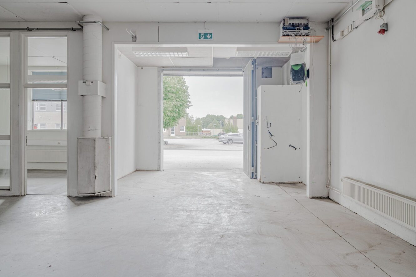 Tomt kommersiellt utrymme med vita väggar och öppen garageport mot en gata som släpper in naturligt ljus; vissa ledningar och armaturer syns på väggarna.