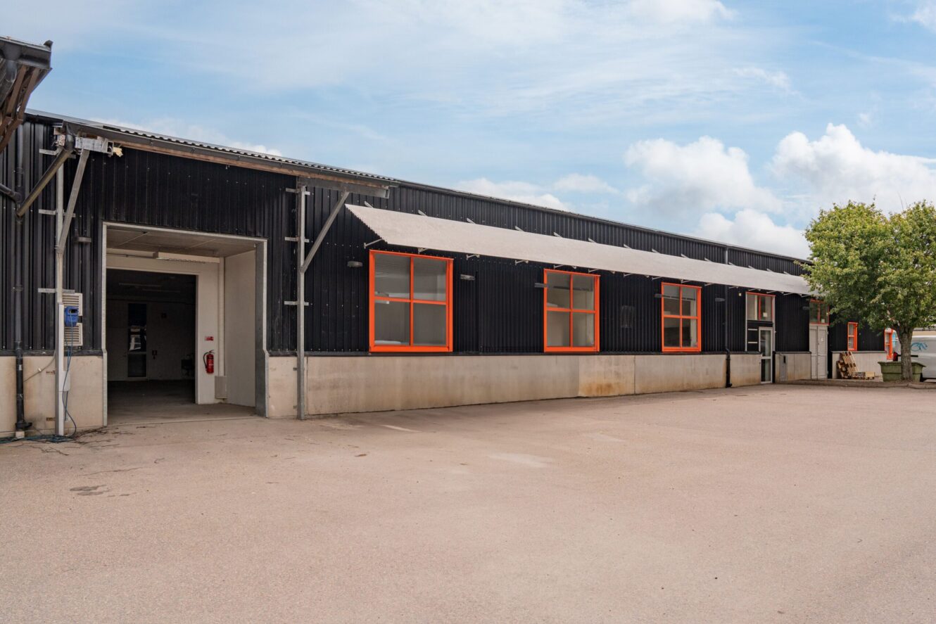 En svart industribyggnad med orangefärgade fönster, betongfundament och en öppen garageport, belägen på en asfalterad tomt under en delvis molnig himmel.