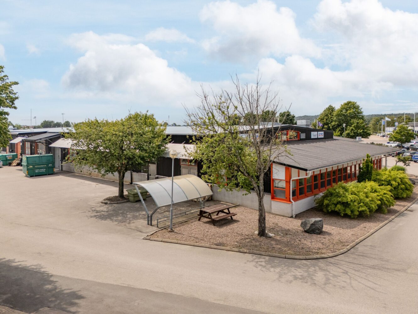 Envåningsbyggnad med orangefärgade fönster, träd, picknickbord, cykelskydd och stora gröna soptunnor på en asfalterad yta under en delvis molnig himmel.