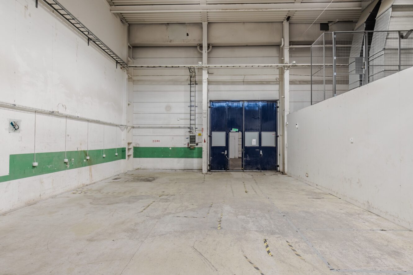 Tomt industriellt lagerutrymme med högt i tak, vita och gröna väggar, rörledningar och stora stängda blå metalldörrar i bortre änden.