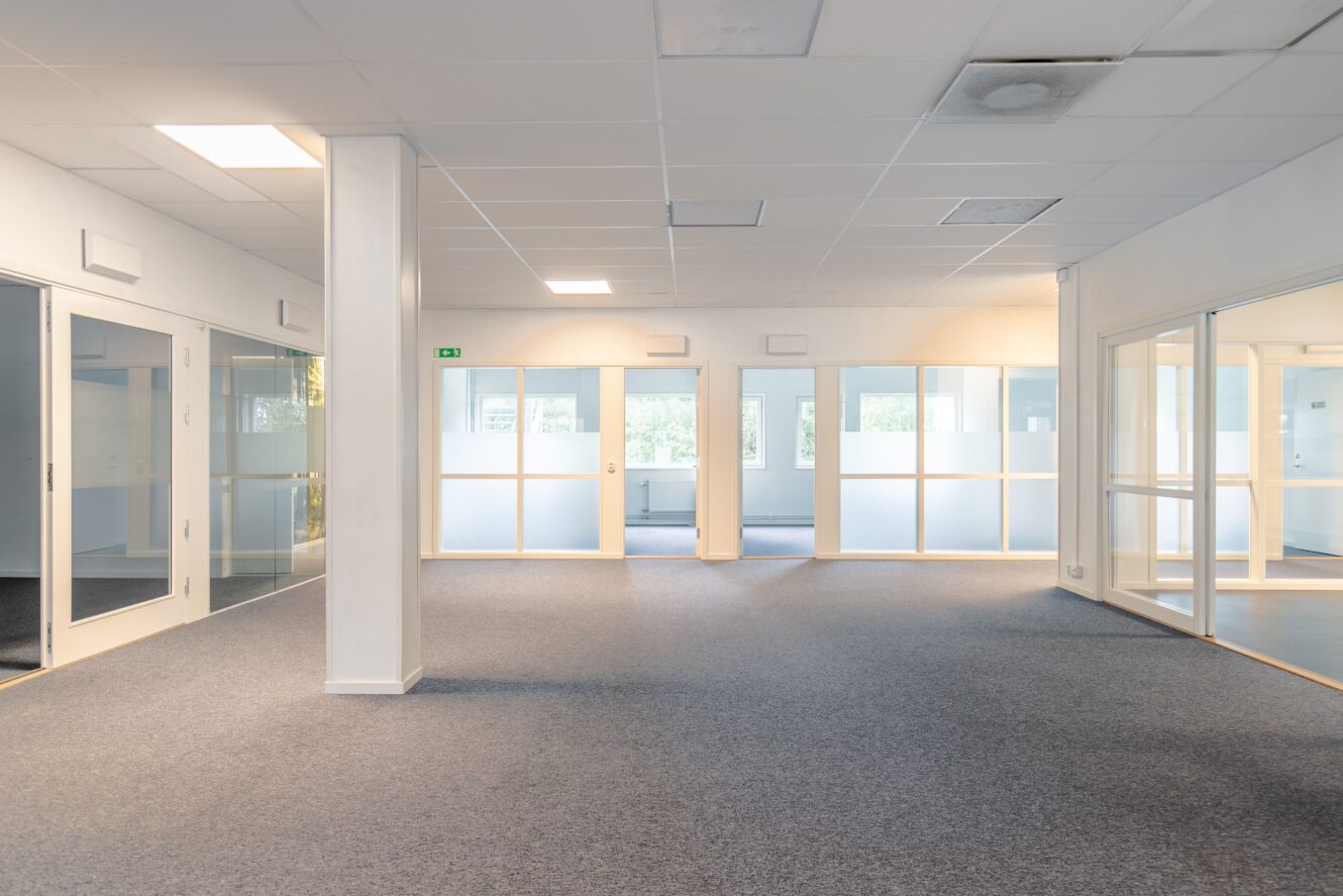 Rymligt, tomt kontor med heltäckningsmatta, glasväggar, vita väggar och lysrör i taket. Naturligt ljus kommer in genom stora fönster och glasdörrar.