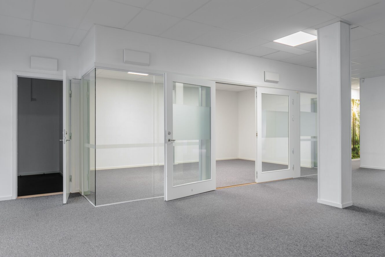 Modern tom kontorslokal med mötesrum med glasväggar, grå matta, vita väggar och taklampor.