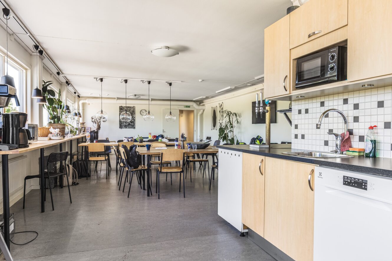 Ett gemensamt kök och matplats med träskåp, vitvaror, flera bord och stolar, stora fönster och växter längs fönsterbrädan.