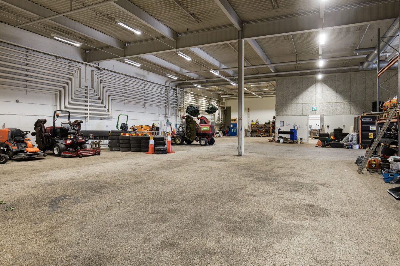 Stort inomhusgarage med olika gräsklippare, utrustning och verktyg längs väggarna; fluorescerande belysning och betonggolv; orangefärgade säkerhetskonor nära mitten.