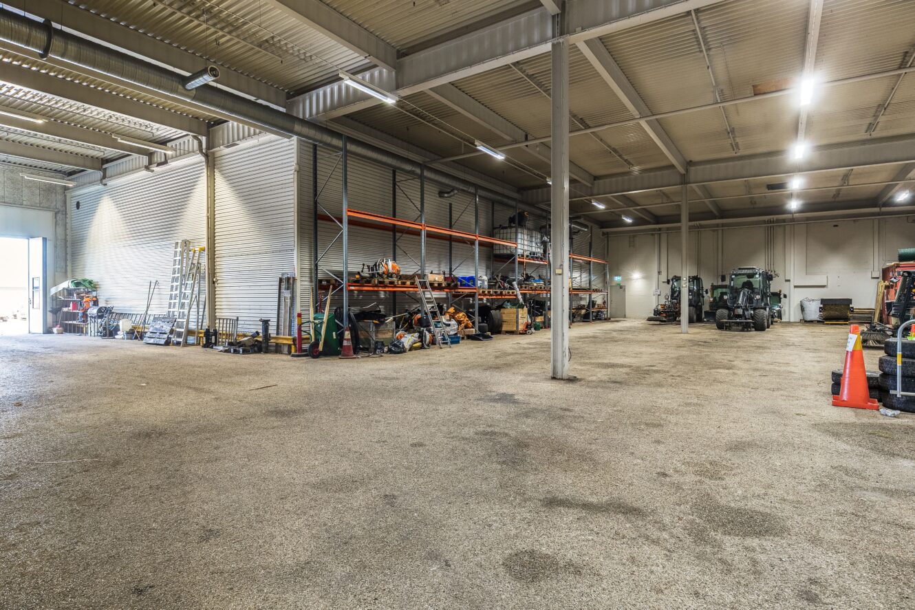 Stort industrilager med betonggolv, metallhyllor med verktyg och utrustning, en gaffeltruck och byggkonor; öppen garagedörr släpper in dagsljus.