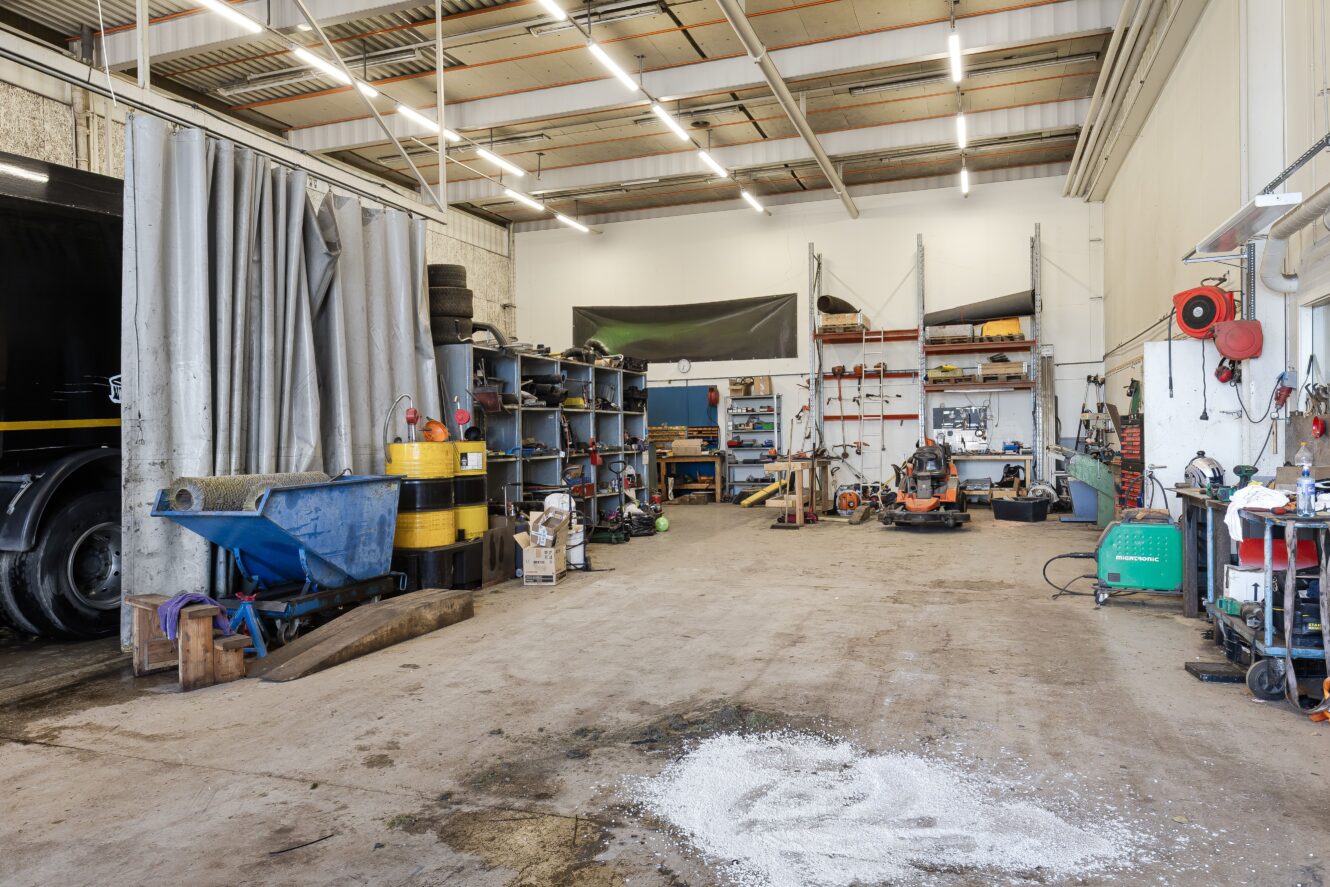 En rymlig verkstad med betonggolv, hyllor med verktyg och förnödenheter, maskiner och diverse utrustning placerade längs väggarna under starka taklampor.