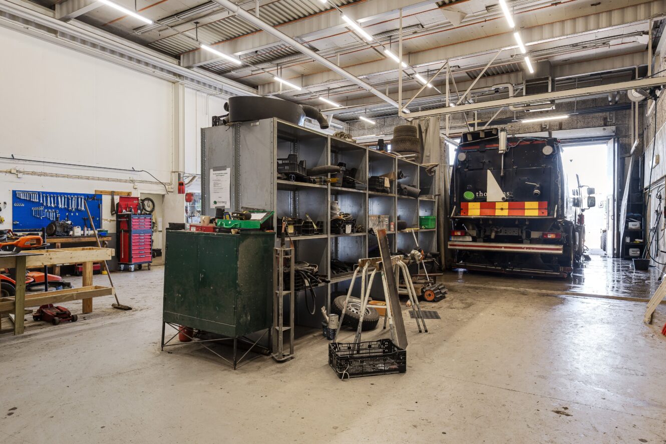 En väl upplyst industriverkstad med organiserade hyllor, arbetsbänkar, verktyg och ett stort bruksfordon parkerat nära den öppna garageporten.