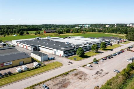 Flygfoto över en stor industribyggnad med parkeringsplats, omgiven av grönska och några andra byggnader under en klar himmel.