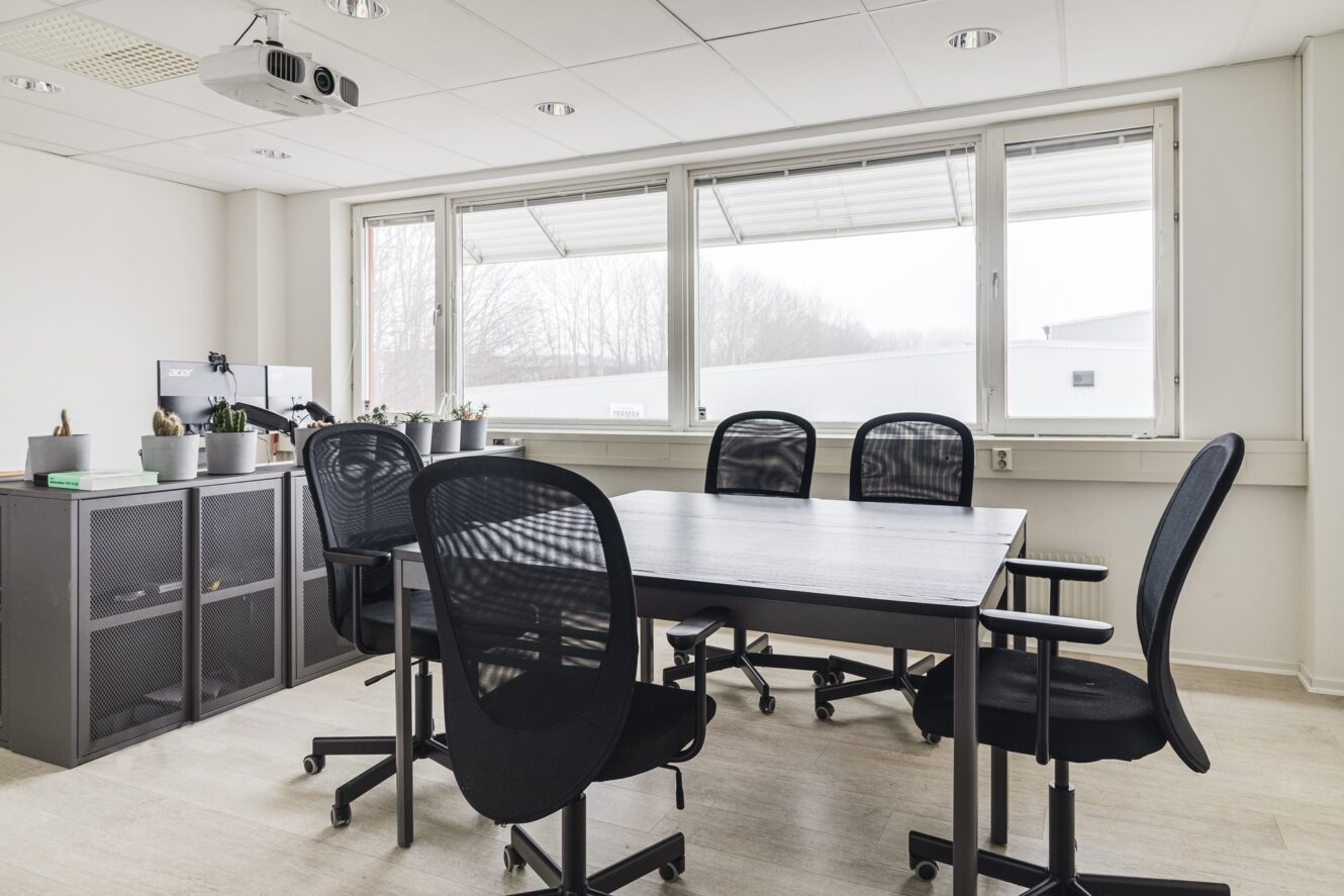 Litet modernt konferensrum med ett rektangulärt bord, fem kontorsstolar i svart mesh, en projektor i taket, stora fönster och krukväxter på ett skåp.