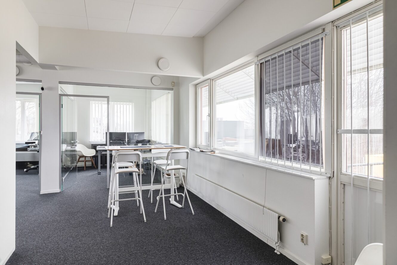 En modern kontorslokal med mötesrum med glasväggar, höga bord och stolar, stora fönster med persienner och inredning i neutrala färger.