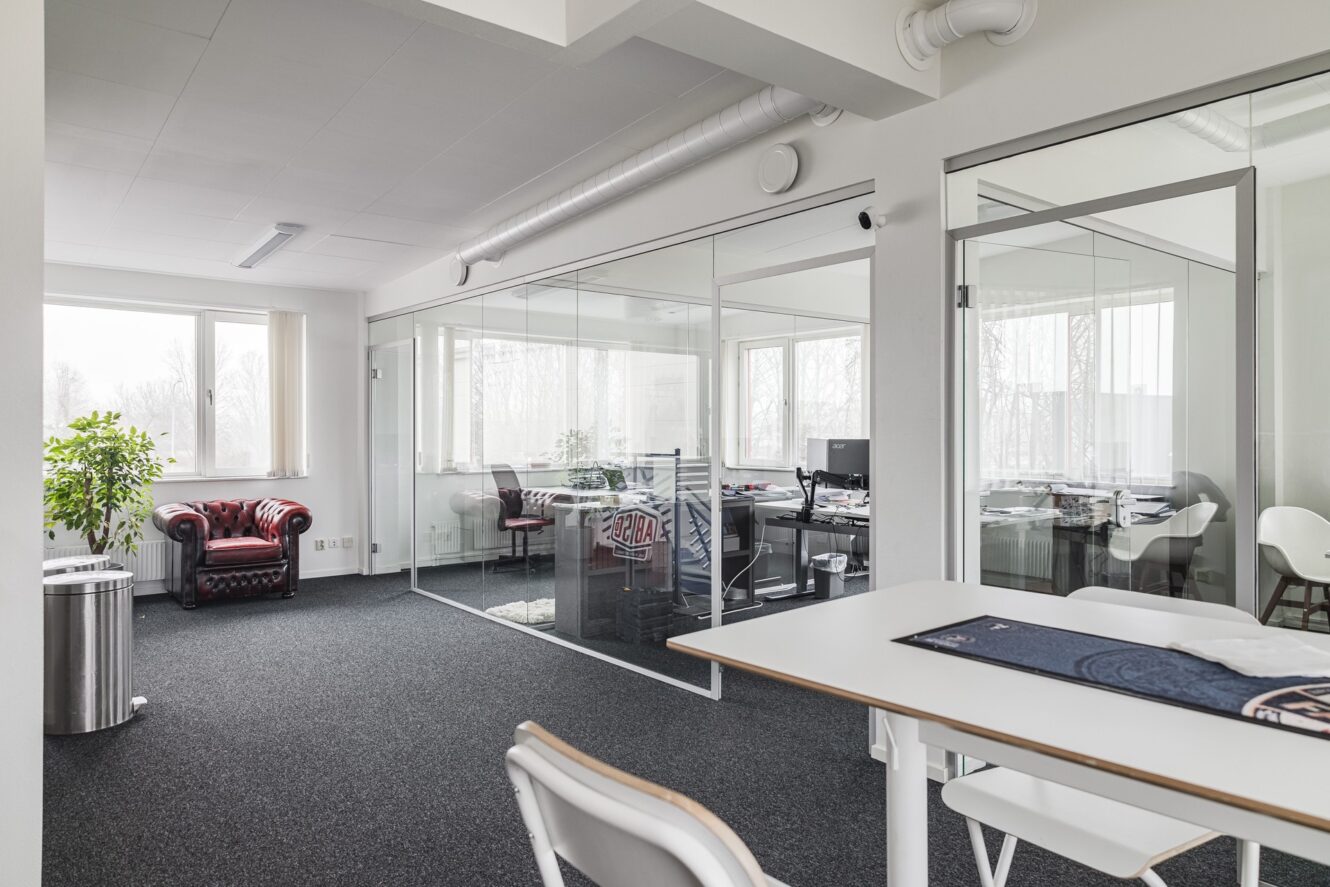 Moderna kontorslokaler med mötesrum med glasväggar, skrivbord med datorer, ett vitt bord med stolar, en krukväxt och en röd fåtölj vid fönstret.
