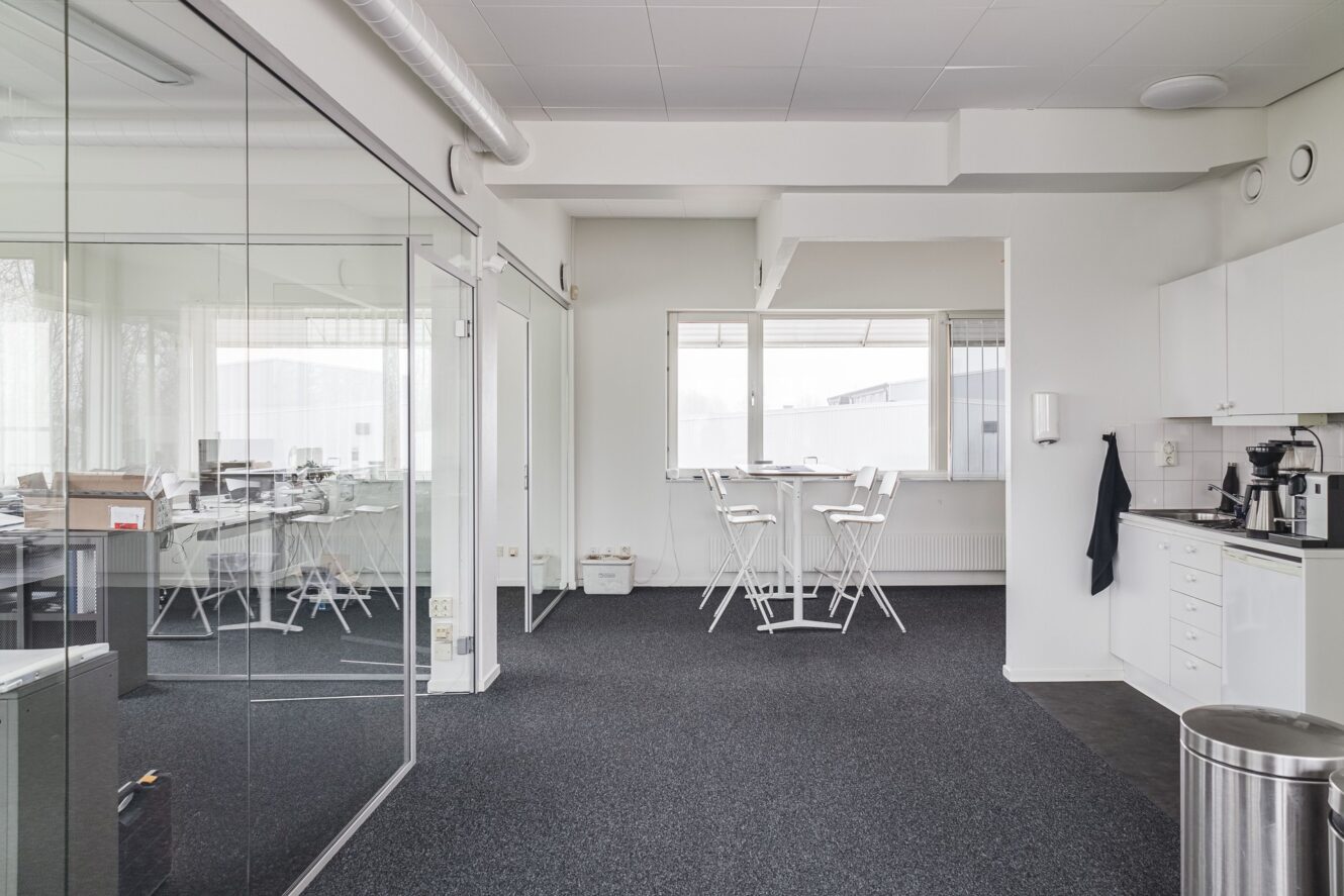 Modernt kontorskök med vita skåp, kaffemaskiner och höga bord intill ett konferensrum med glasväggar, stolar och bord.