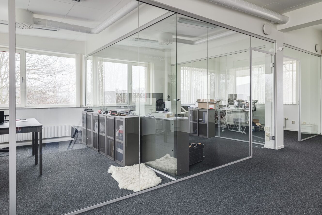 Moderna kontorslokaler med glasväggar, skrivbord, kontorsstolar, hyllor och stora fönster som släpper in naturligt ljus.