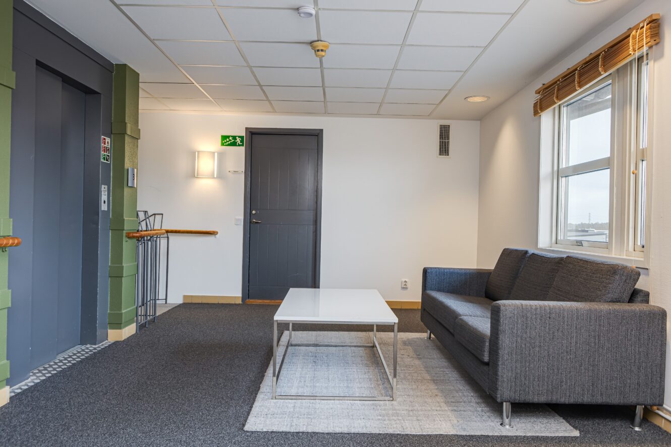 En modern hall med en grå soffa, ett vitt soffbord, en heltäckningsmatta, ett fönster med persienner, en hiss och en stängd grå dörr under en utgångsskylt.