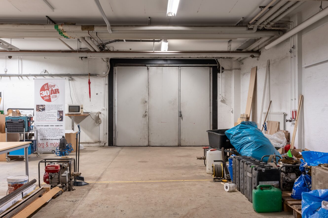 Invändigt industriellt garage med stängda dubbeldörrar i metall, betonggolv, diverse verktyg, utrustning och förnödenheter utspridda runt omkring, inklusive en blå presenning som täcker föremålen på höger sida.