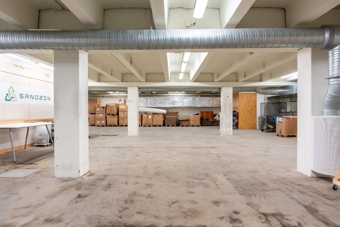 Stor, tom lagerlokal med betonggolv, vitt tak, synliga kanaler, pelare och staplade kartonger mot bakväggen.